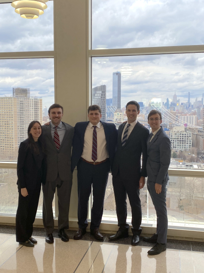 four men and one woman in business suits standing next to each other and smiling in front of large windows with cityscape in distance